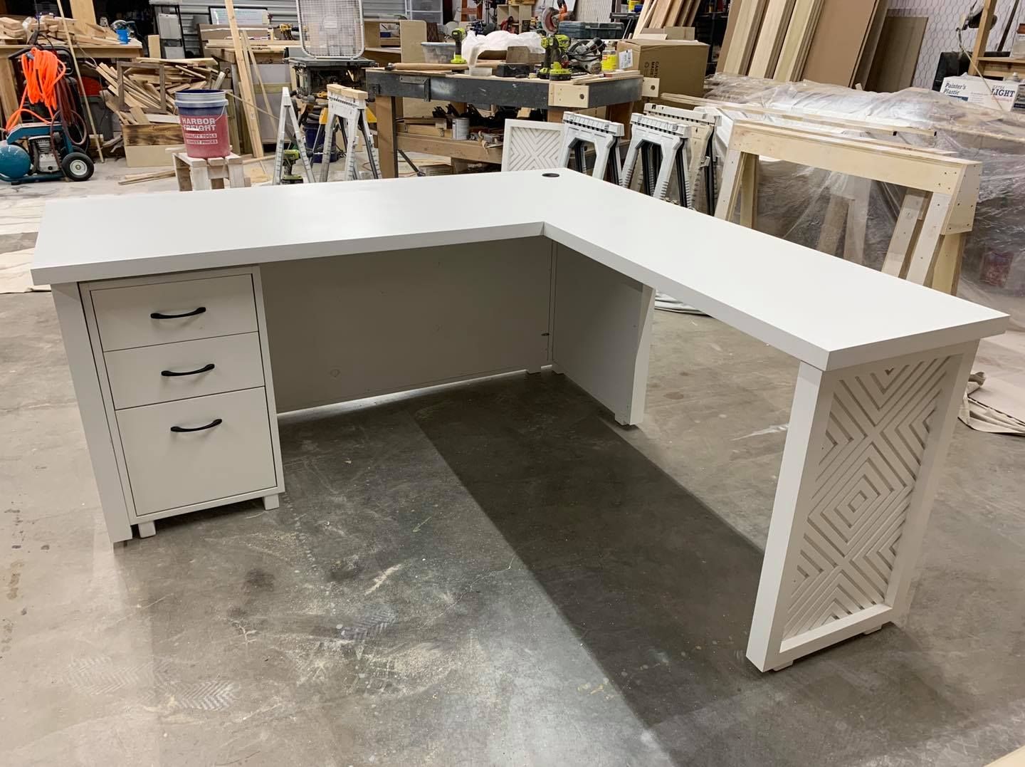 White L-shaped desk with drawers and decorative side panel in a workshop.