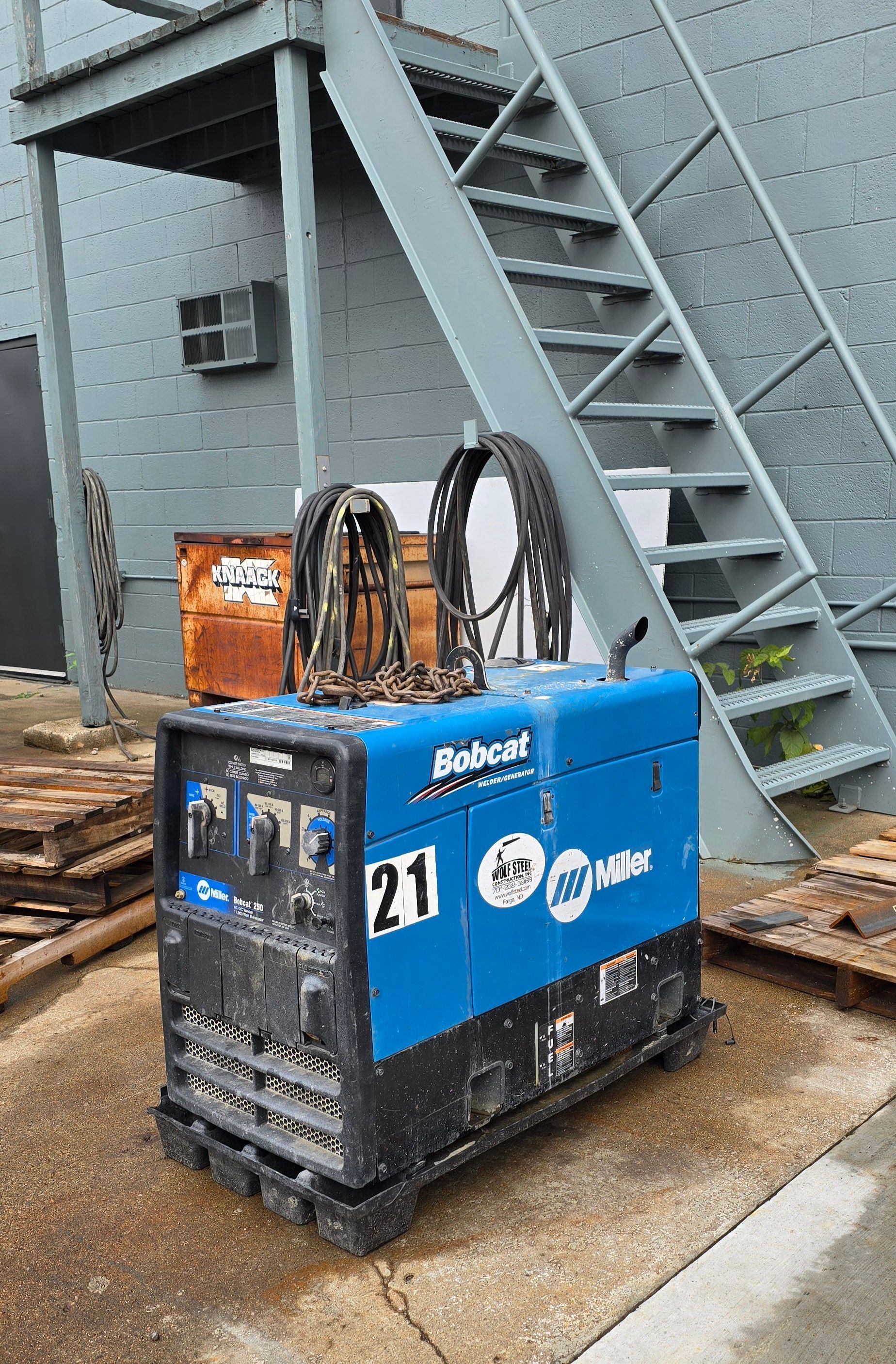 A welding machine is parked in front of a building next to a set of stairs.