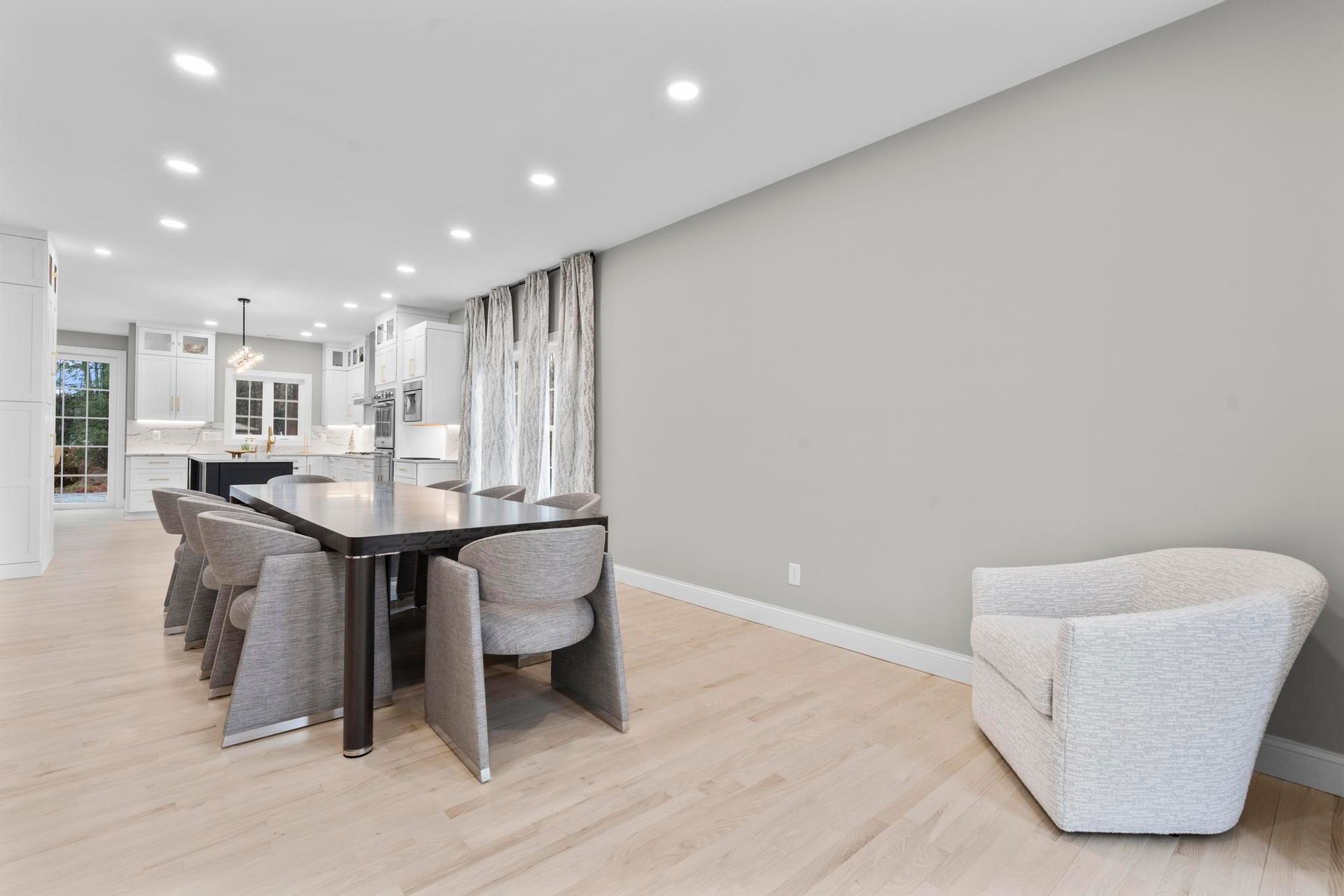 A modern dining area with a long table, grey chairs, light wood floors, and a single white armchair against a grey wall.
