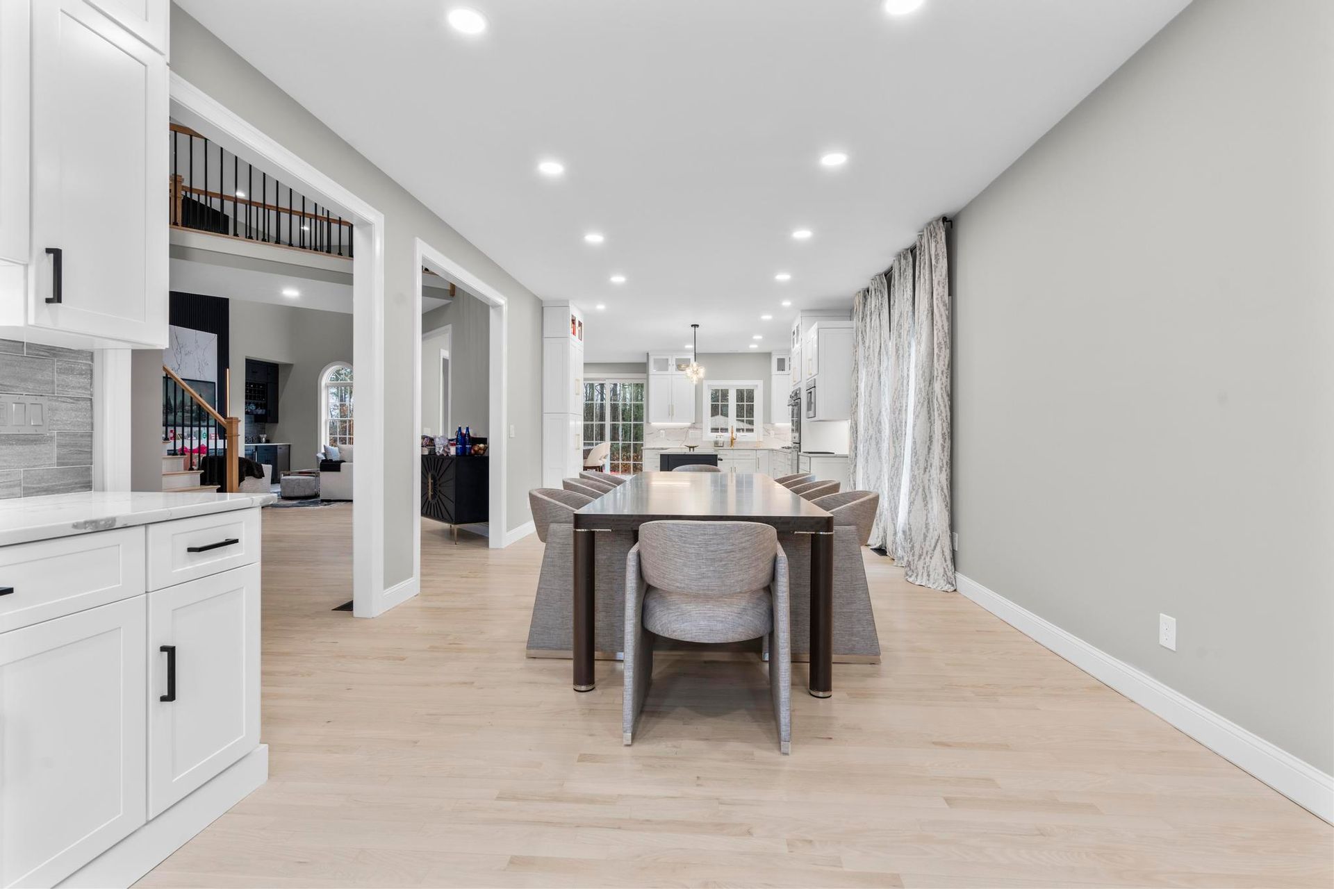 A modern, open-concept dining area with a long wooden table, grey upholstered chairs, light hardwood floors, and curtains.