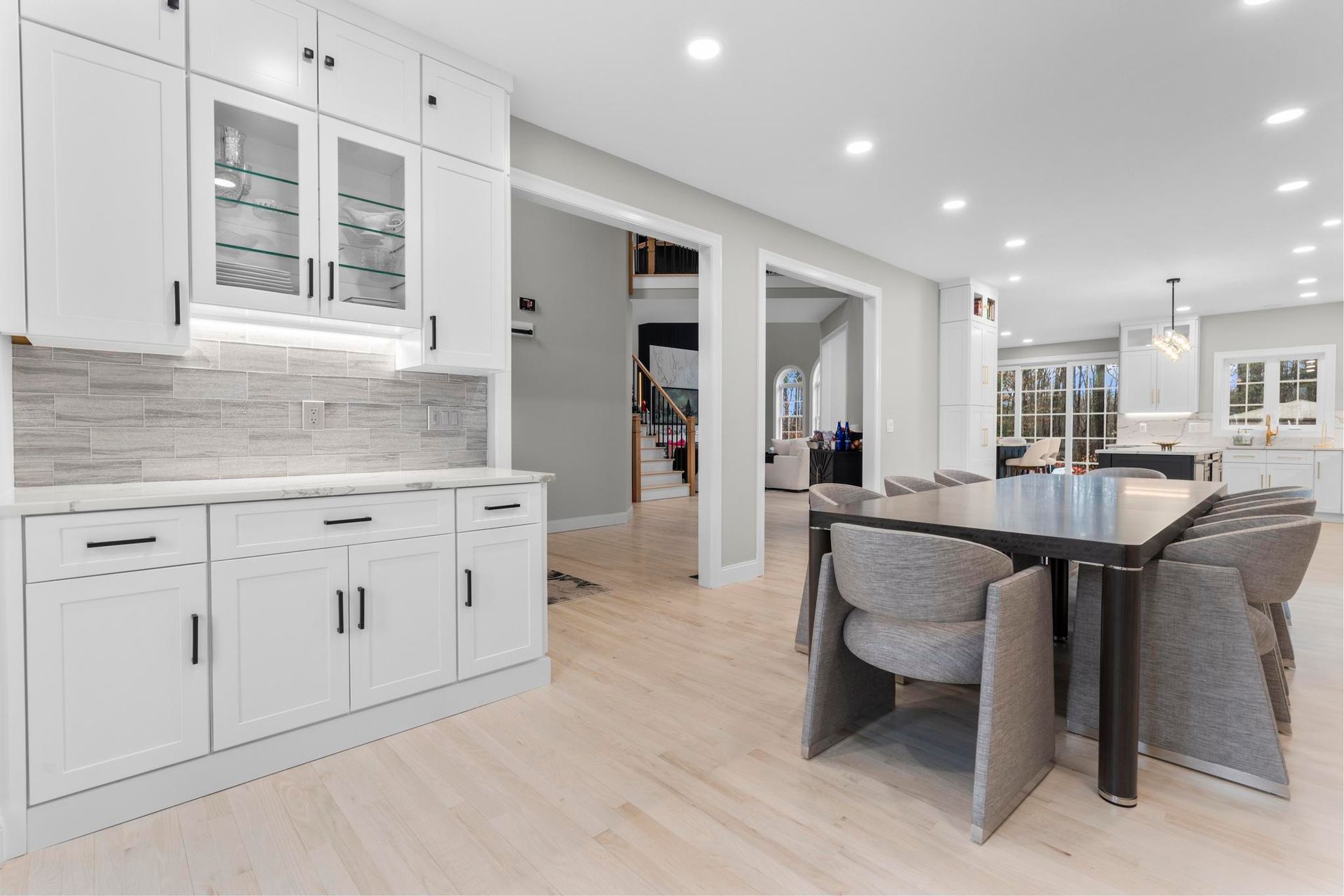A modern kitchen area featuring white cabinetry with glass-front uppers, light hardwood floors, and a dining table.