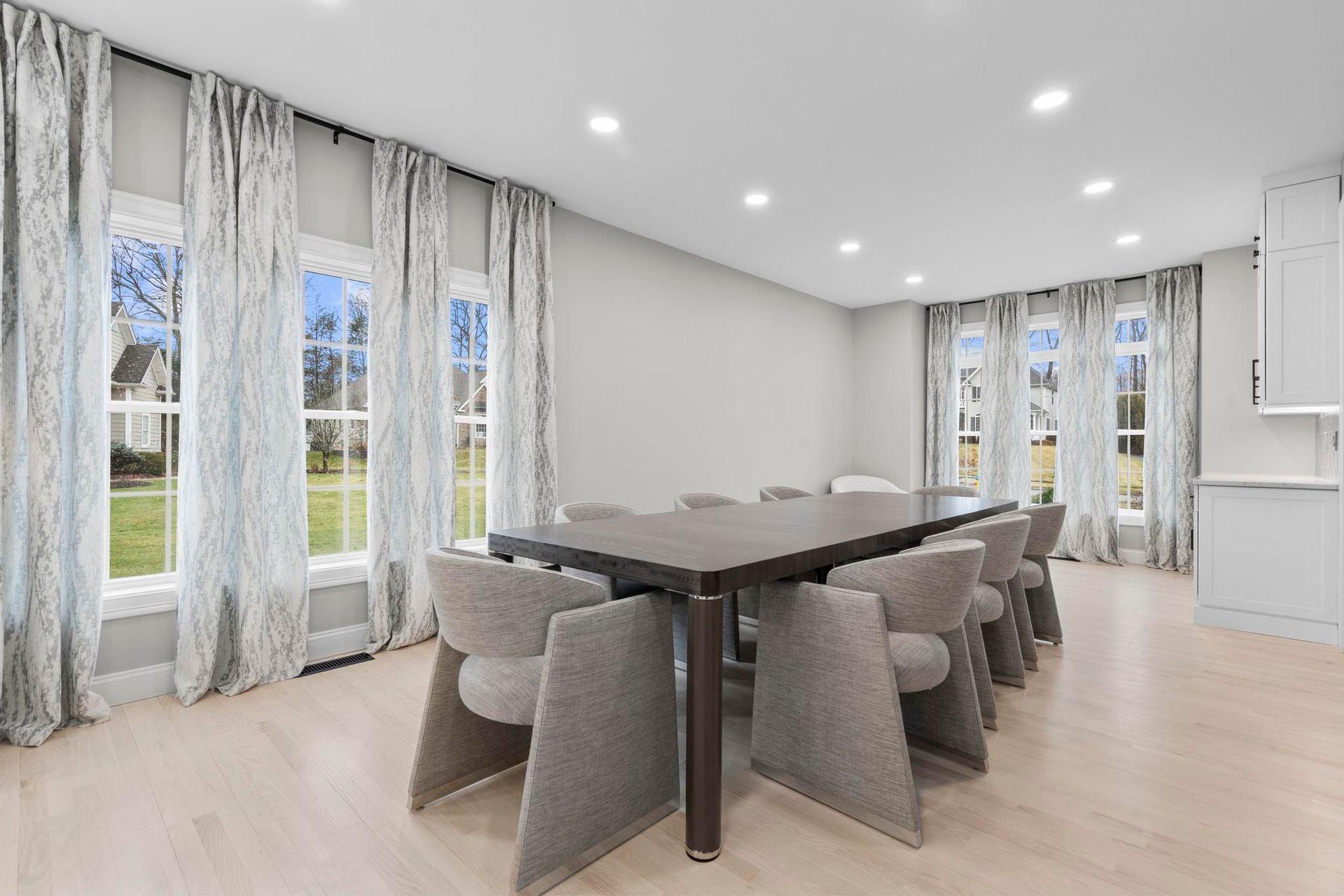 A bright dining room with light wood floors, a dark rectangular table, and grey upholstered chairs under recessed lights.