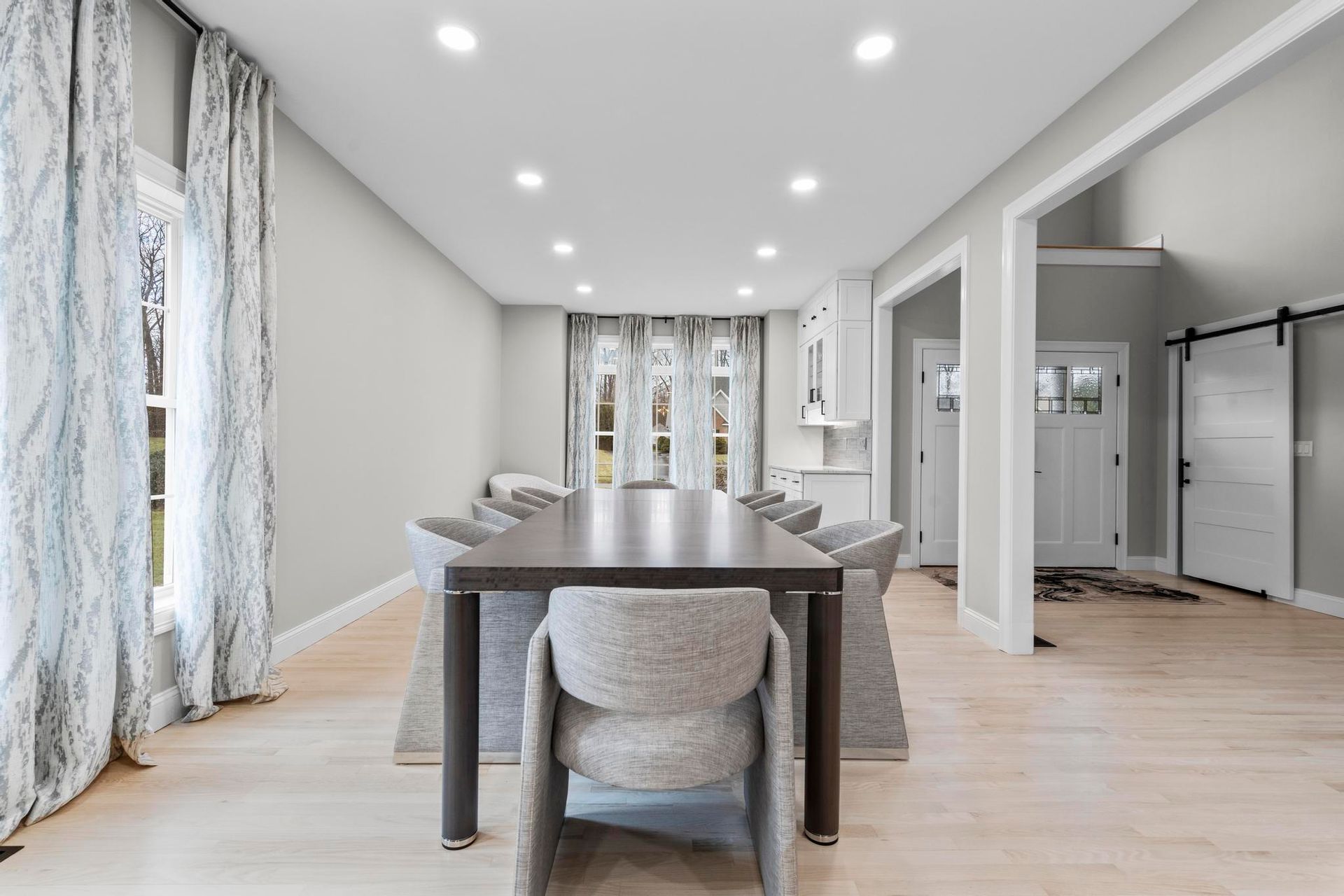 A dining room with a long dark wooden table, upholstered grey chairs, light wood floors, and patterned curtains.