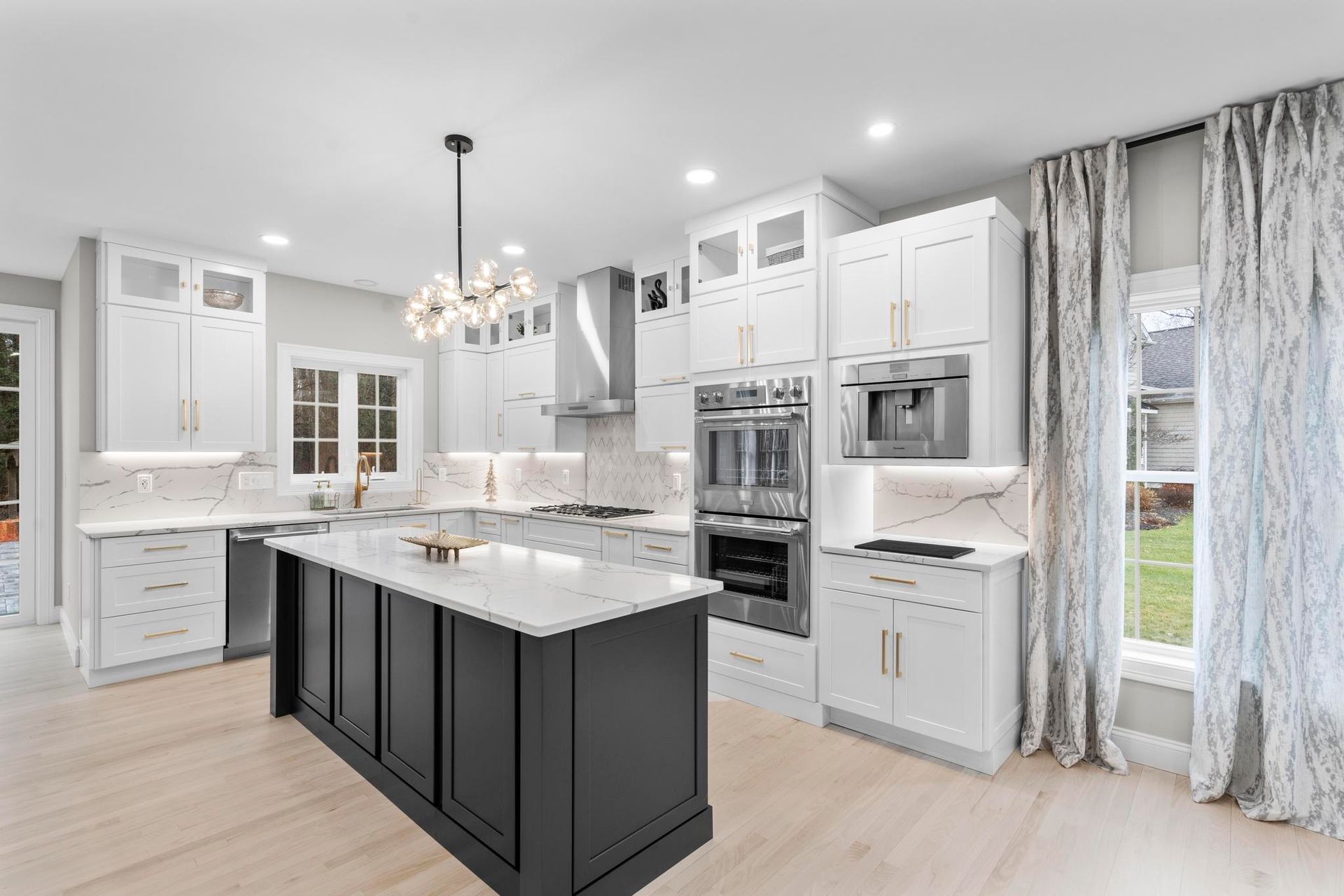A modern kitchen featuring white cabinets, a dark gray island with marble countertops, stainless appliances, and curtains.