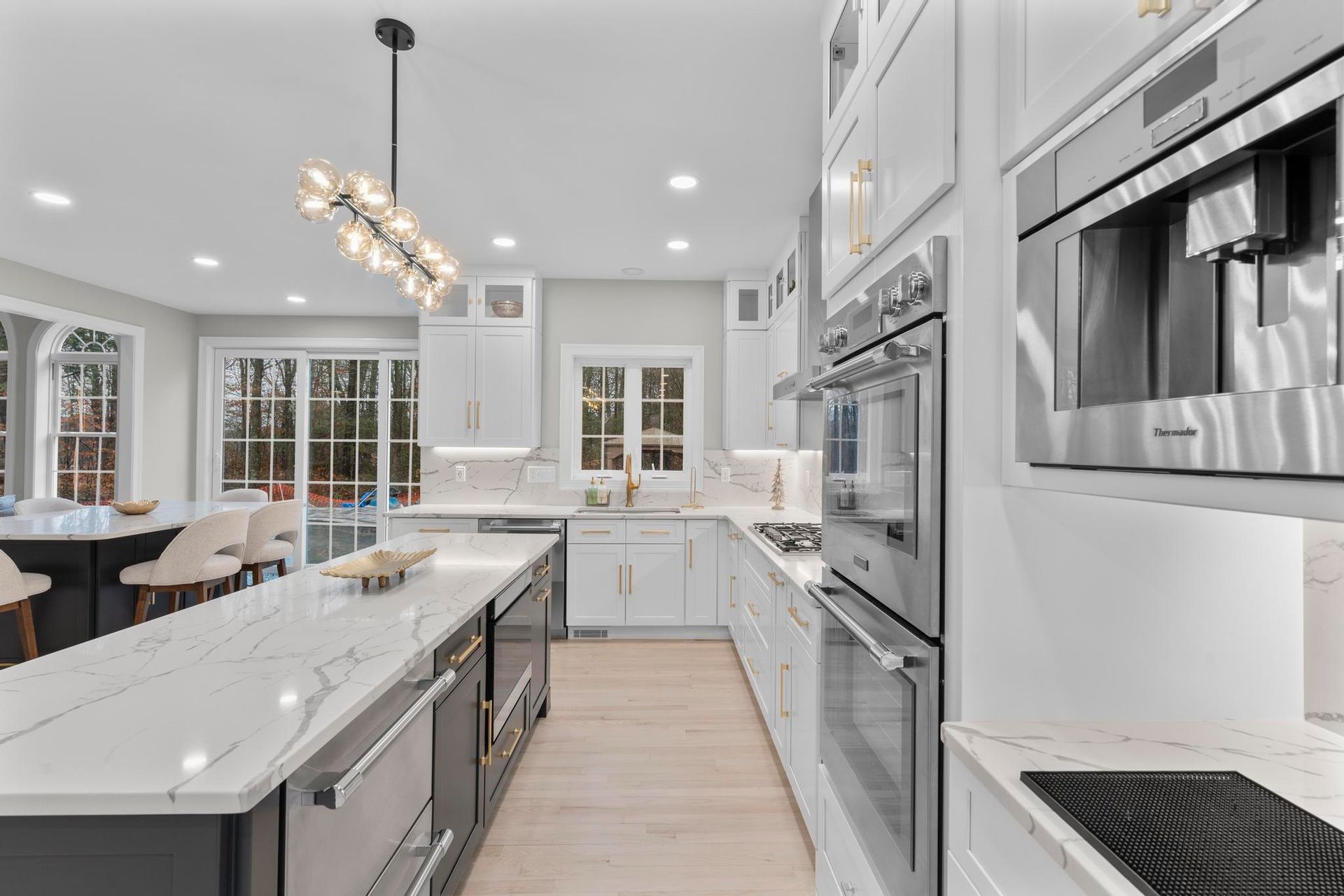 A bright, modern kitchen features a long white marble island, white cabinetry, stainless steel appliances, and a chandelier.