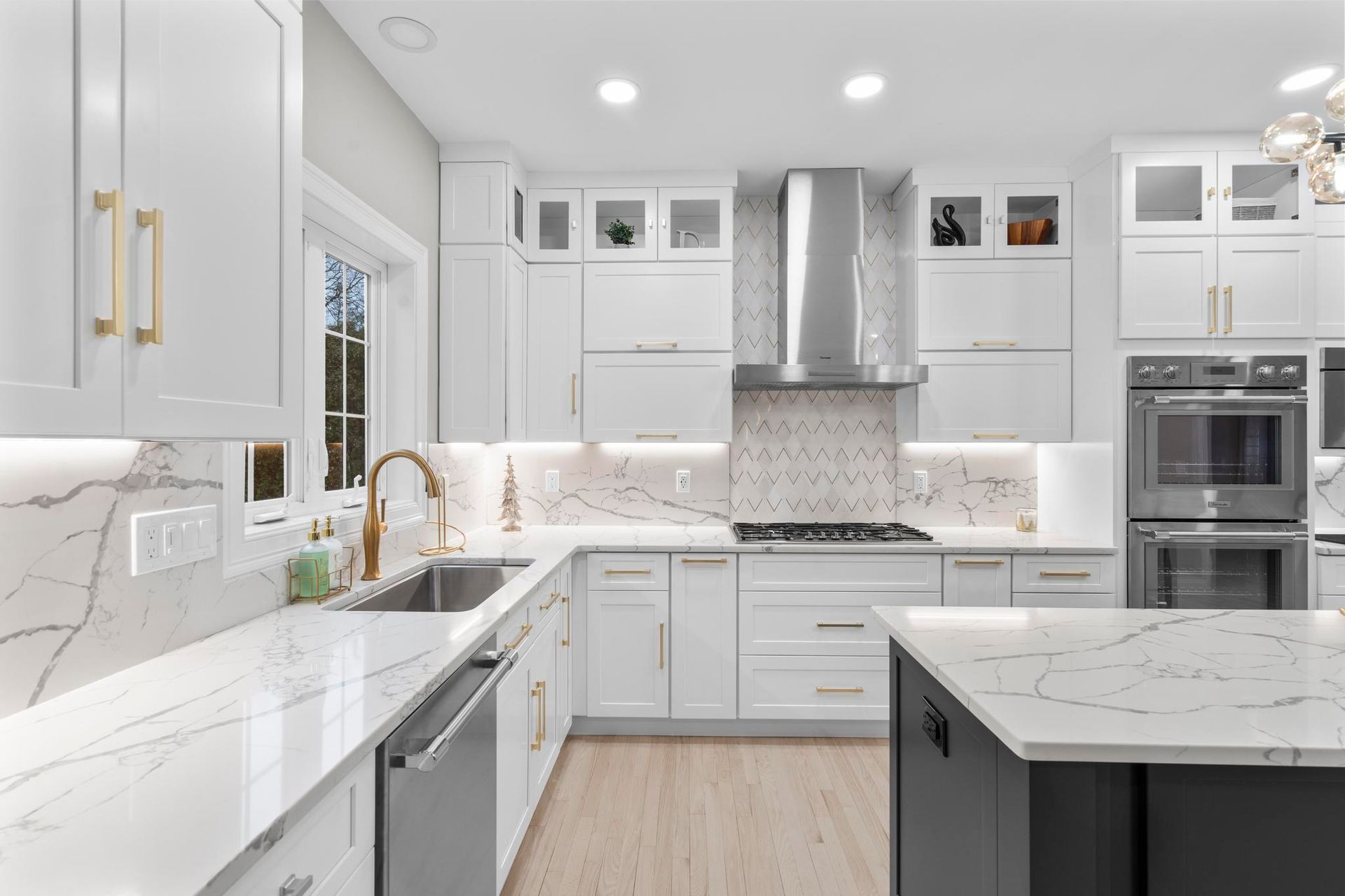 A modern white kitchen featuring marble countertops, a charcoal island, stainless steel appliances, and gold hardware.