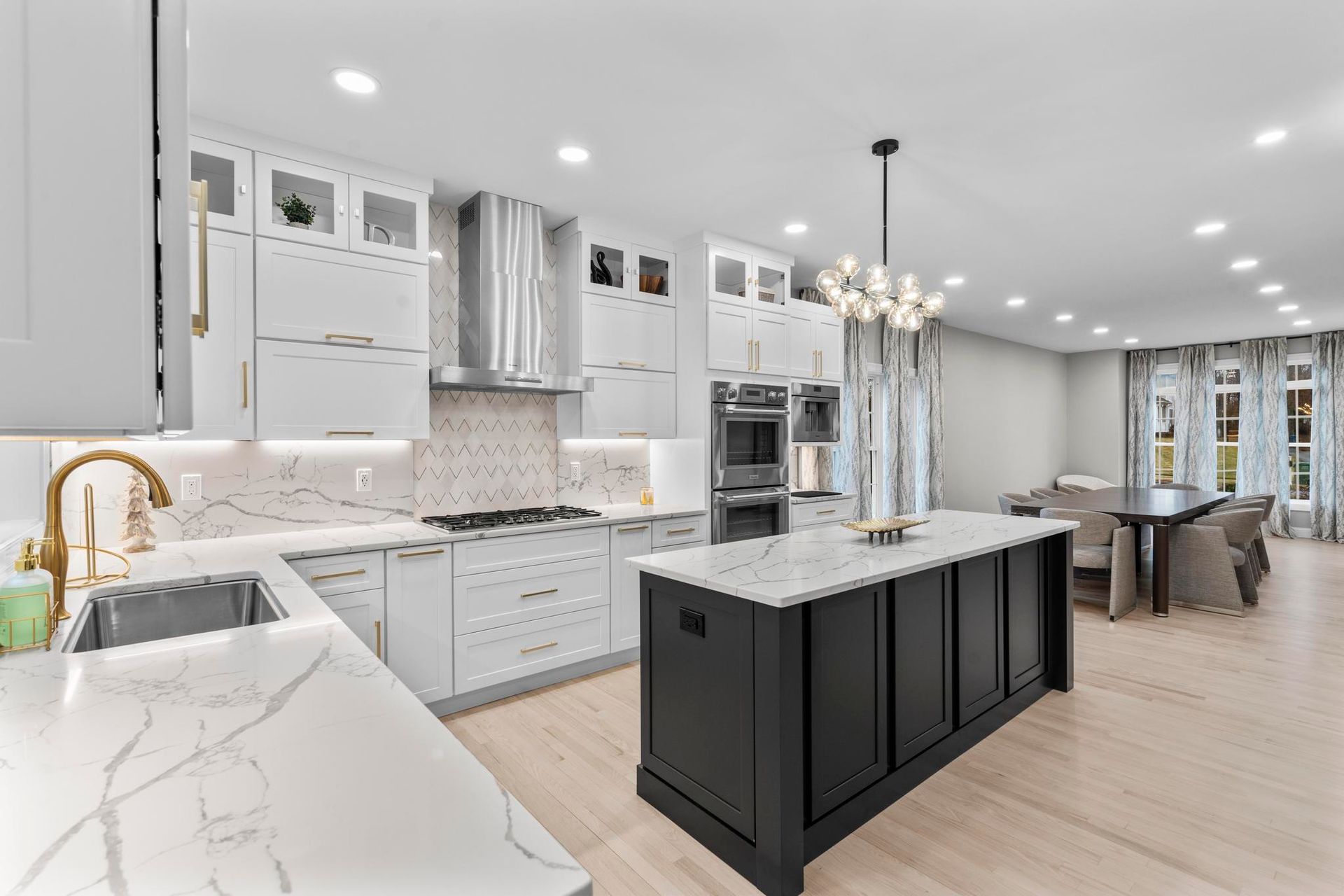 Modern kitchen featuring white cabinets, marble countertops, a black center island, stainless steel appliances, and lighting.