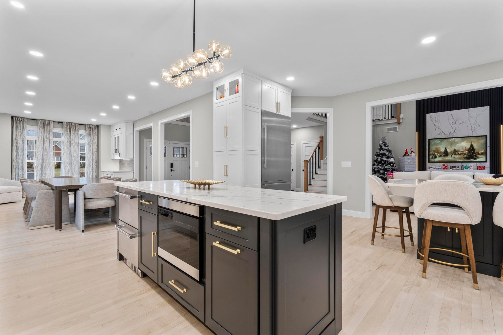 A modern kitchen with a black island, white marble countertops, and light wood floors, opening to a dining area.