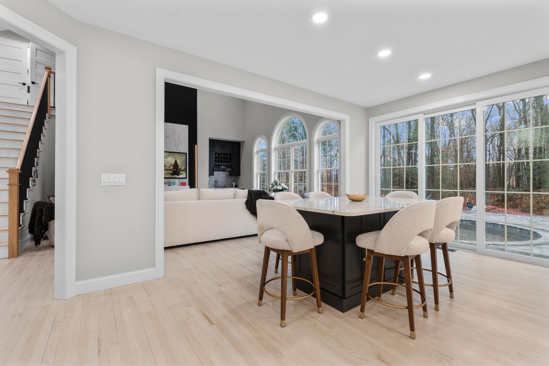 A kitchen island with four cream chairs in a bright, modern room featuring light wood floors and a view of a backyard.
