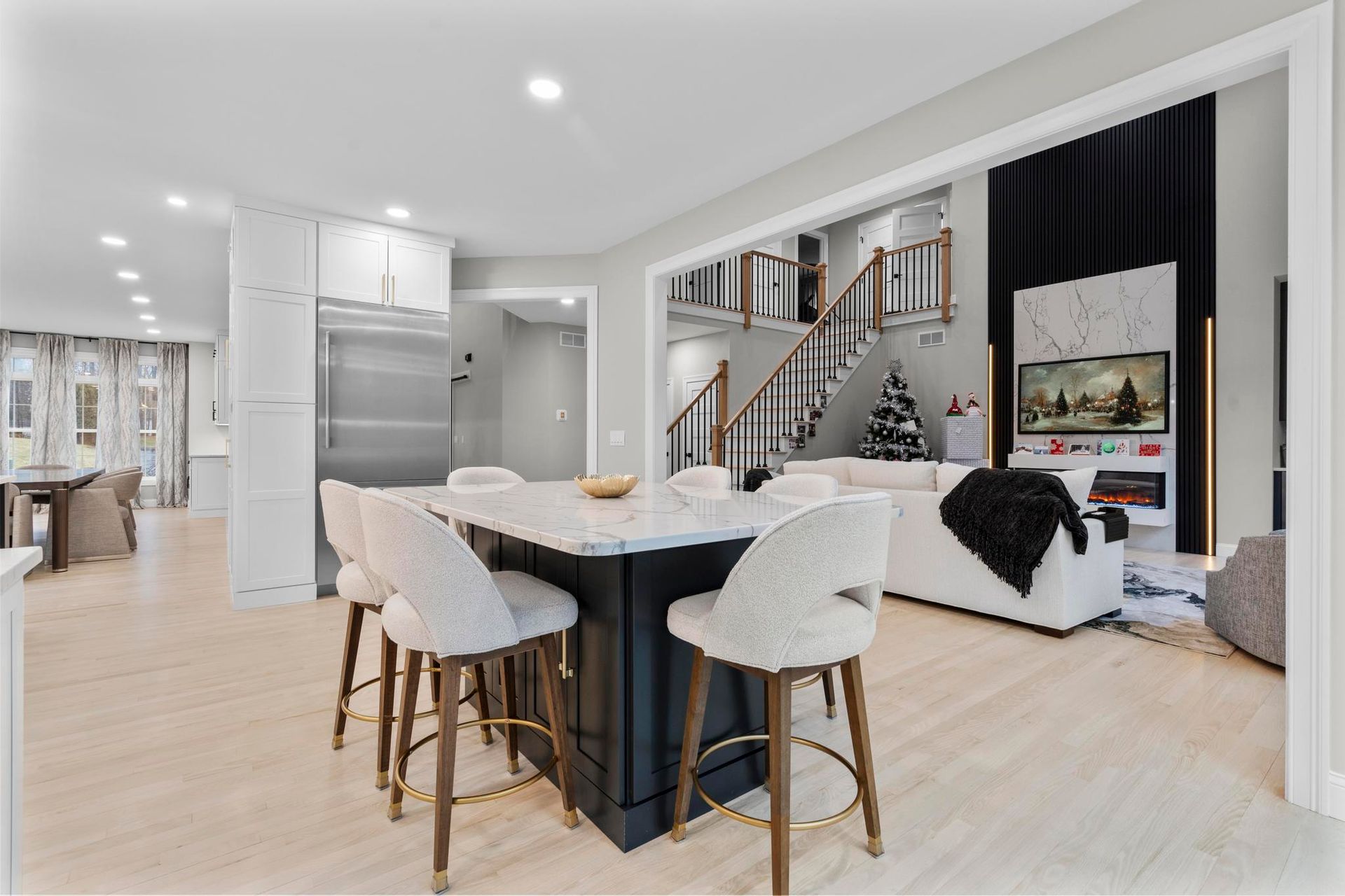 A modern kitchen with a dark blue island and light stools, opening into a spacious living room with a staircase.