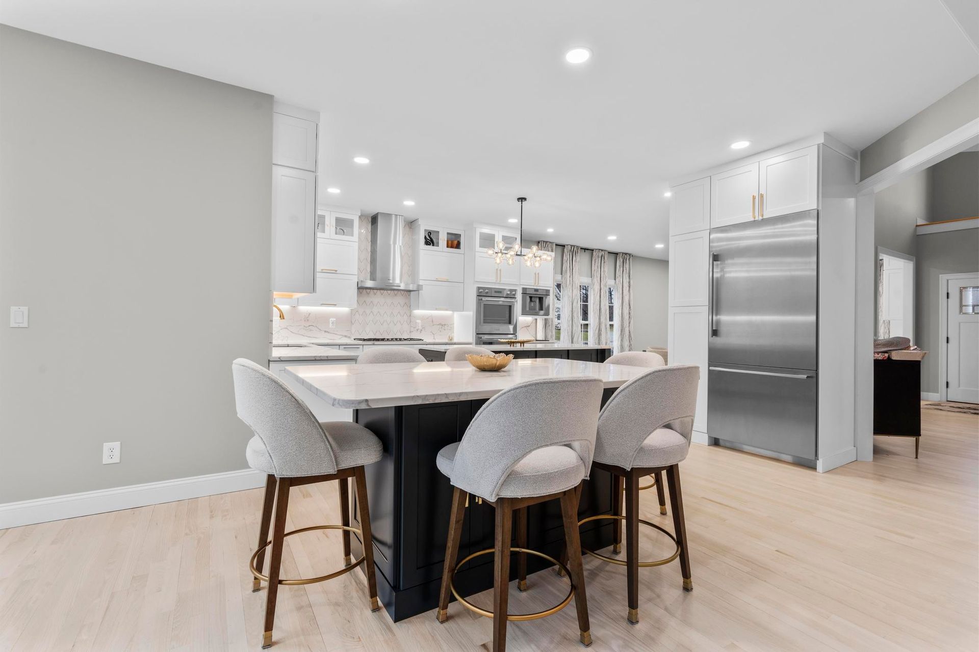 A modern kitchen with a dark island and light stools, stainless steel appliances, and light-colored hardwood floors.