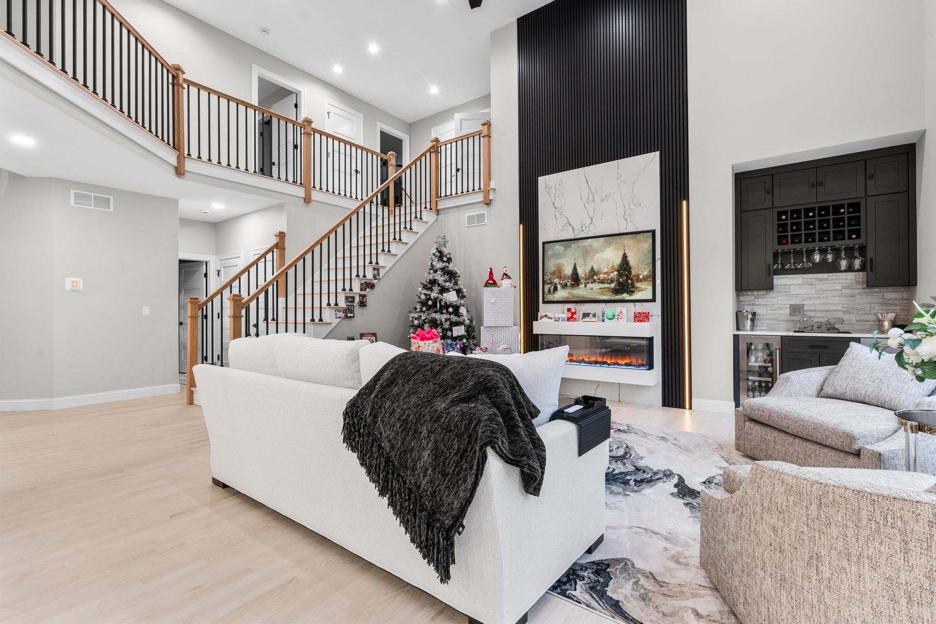 A bright living room with a white sofa, a staircase with wood railings, a fireplace wall, and a built-in home bar.