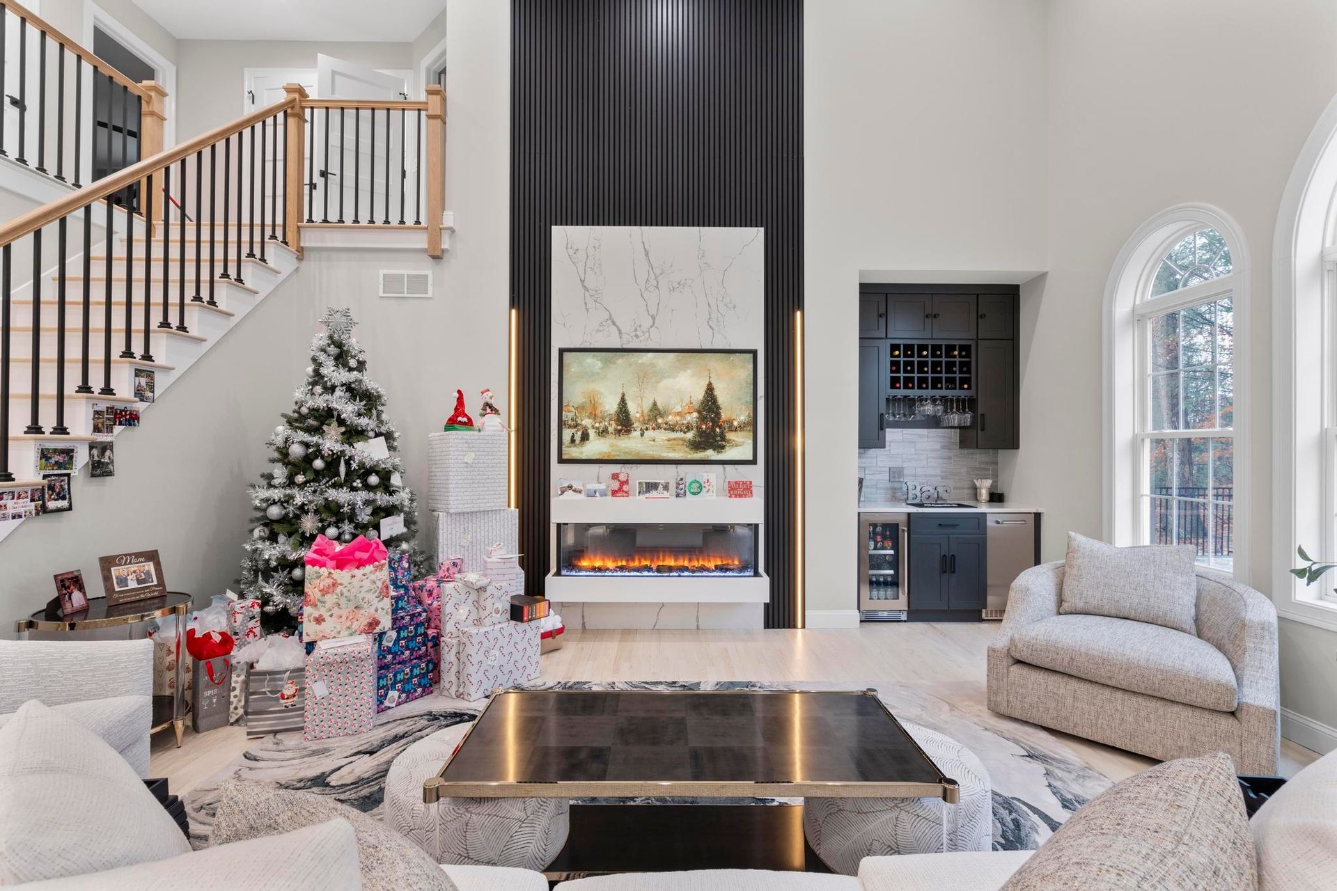 A decorated Christmas tree and gifts sit in a bright, modern living room with a tall fireplace and built-in wet bar.