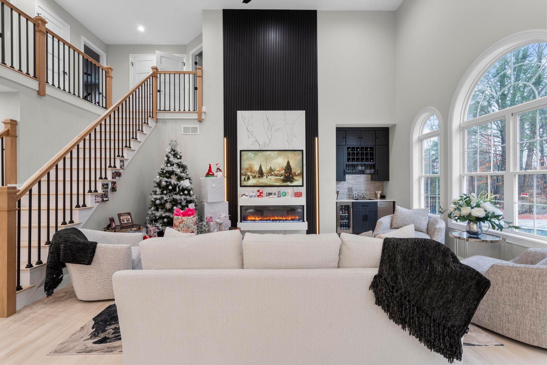 A modern living room with a tall fireplace, a staircase, a Christmas tree, and neutral seating.