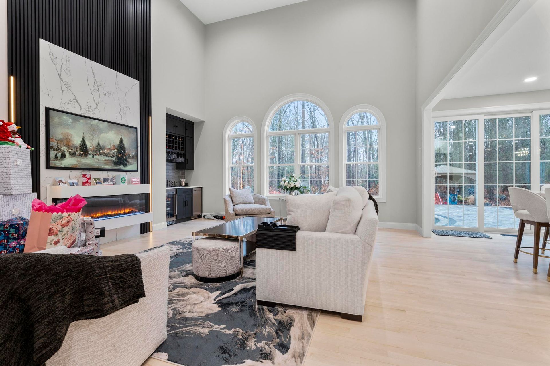A bright, high-ceilinged living room with white furniture, a modern fireplace, large arched windows, and a patterned rug.