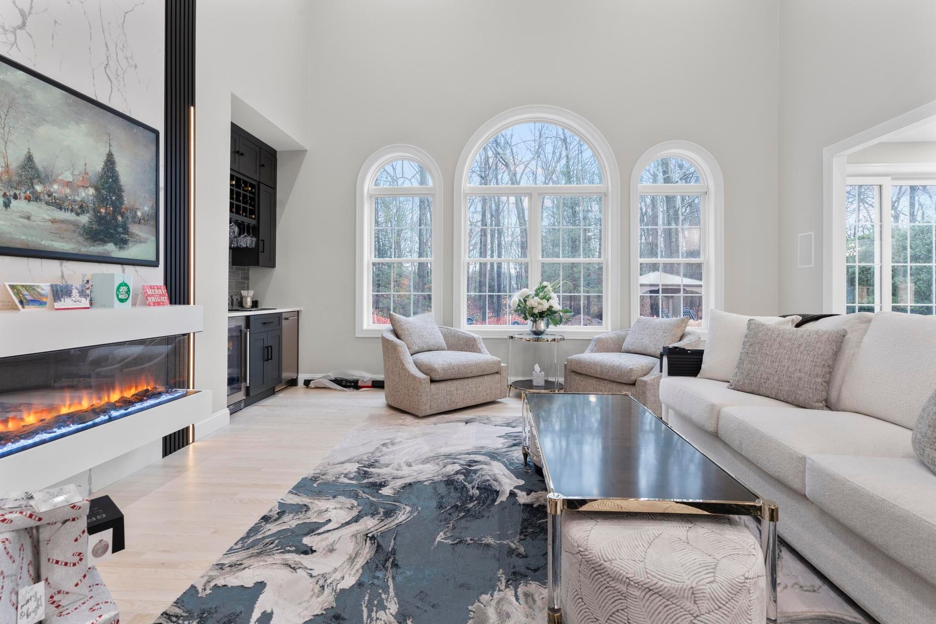 A bright living room with tall windows, a modern fireplace, a sectional sofa, two armchairs, and a patterned rug.