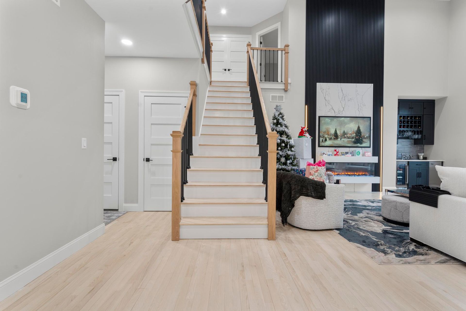 A brightly lit modern home interior featuring a central staircase, light wood floors, white doors, and a living area.