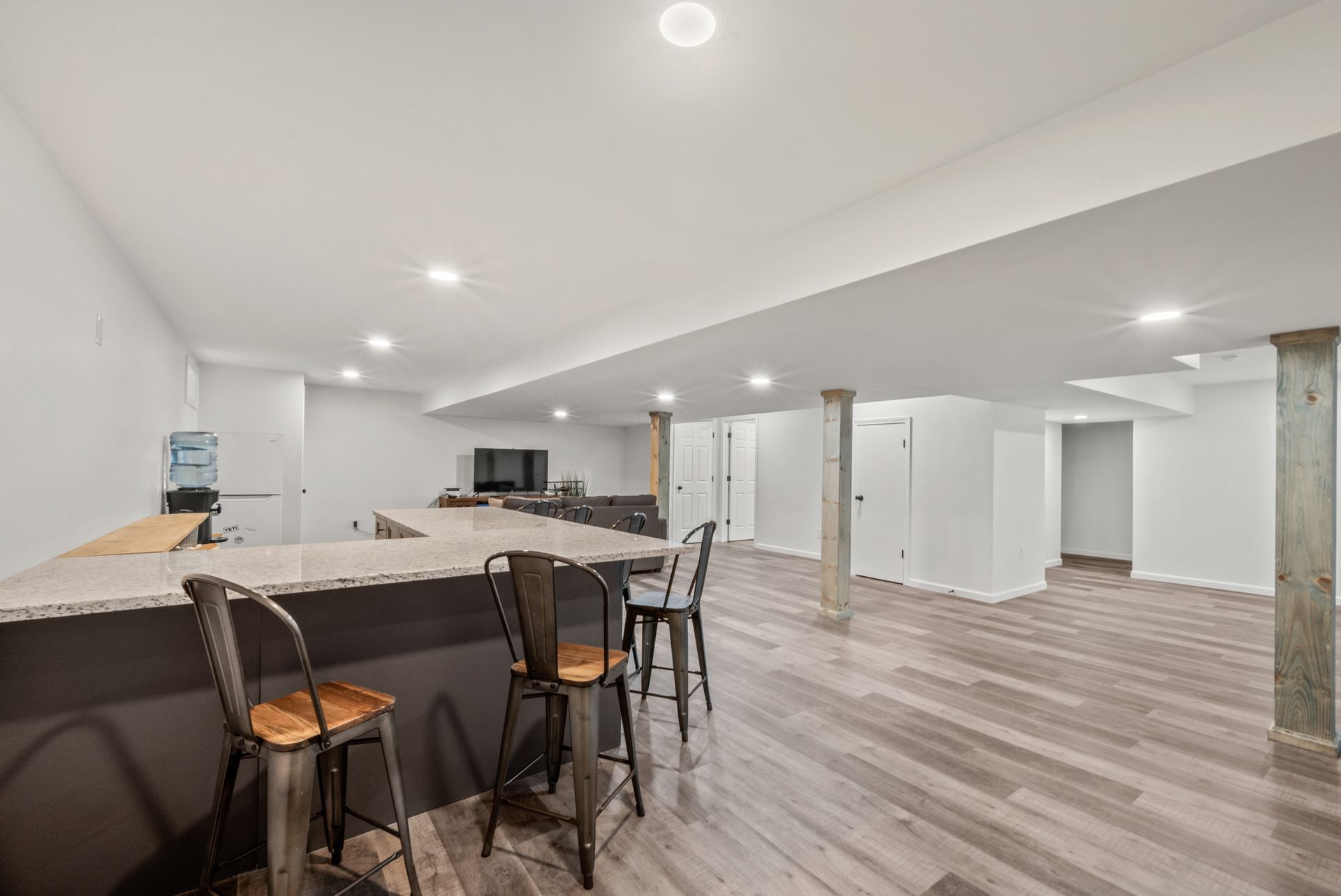 A finished basement with light wood flooring, white walls, recessed lighting, a granite bar, and wooden support columns.