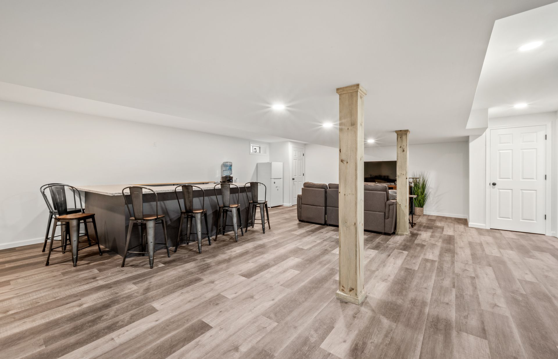 A finished basement with wood flooring, a long bar with stools, two wooden support pillars, and a living area.