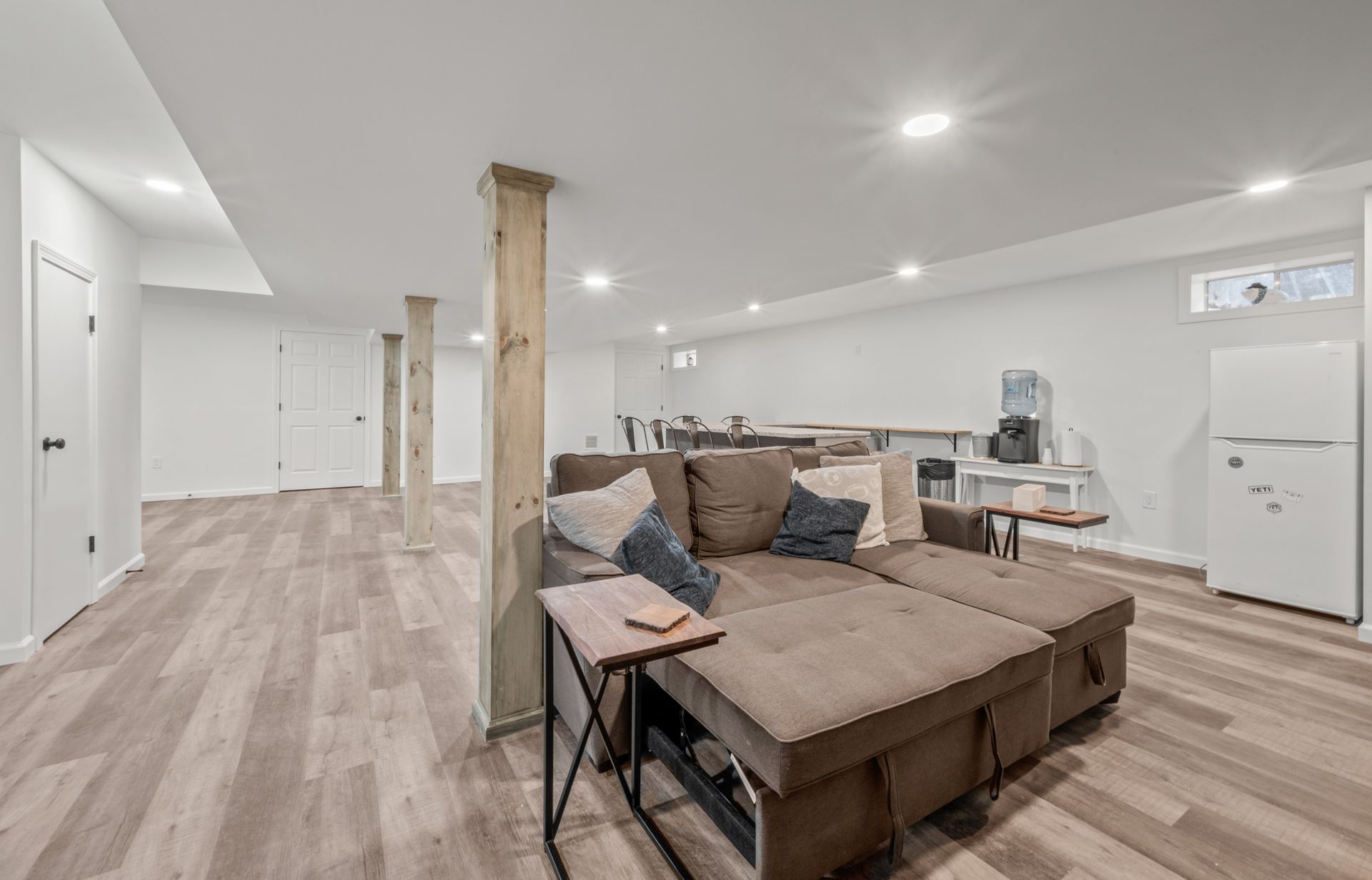 A finished basement featuring light wood-look flooring, a brown sectional sofa, exposed wood support beams, and a mini fridge.