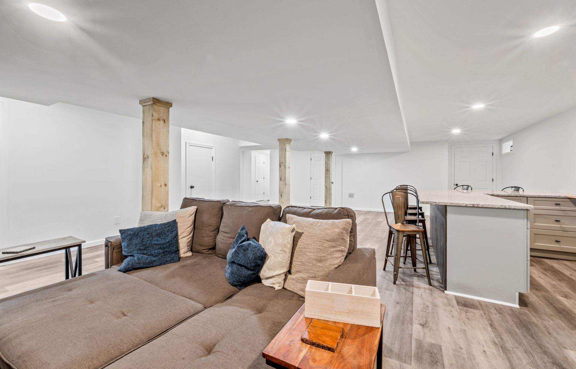 A furnished basement with a large brown sectional, wooden support beams, recessed lighting, and a small kitchen island.