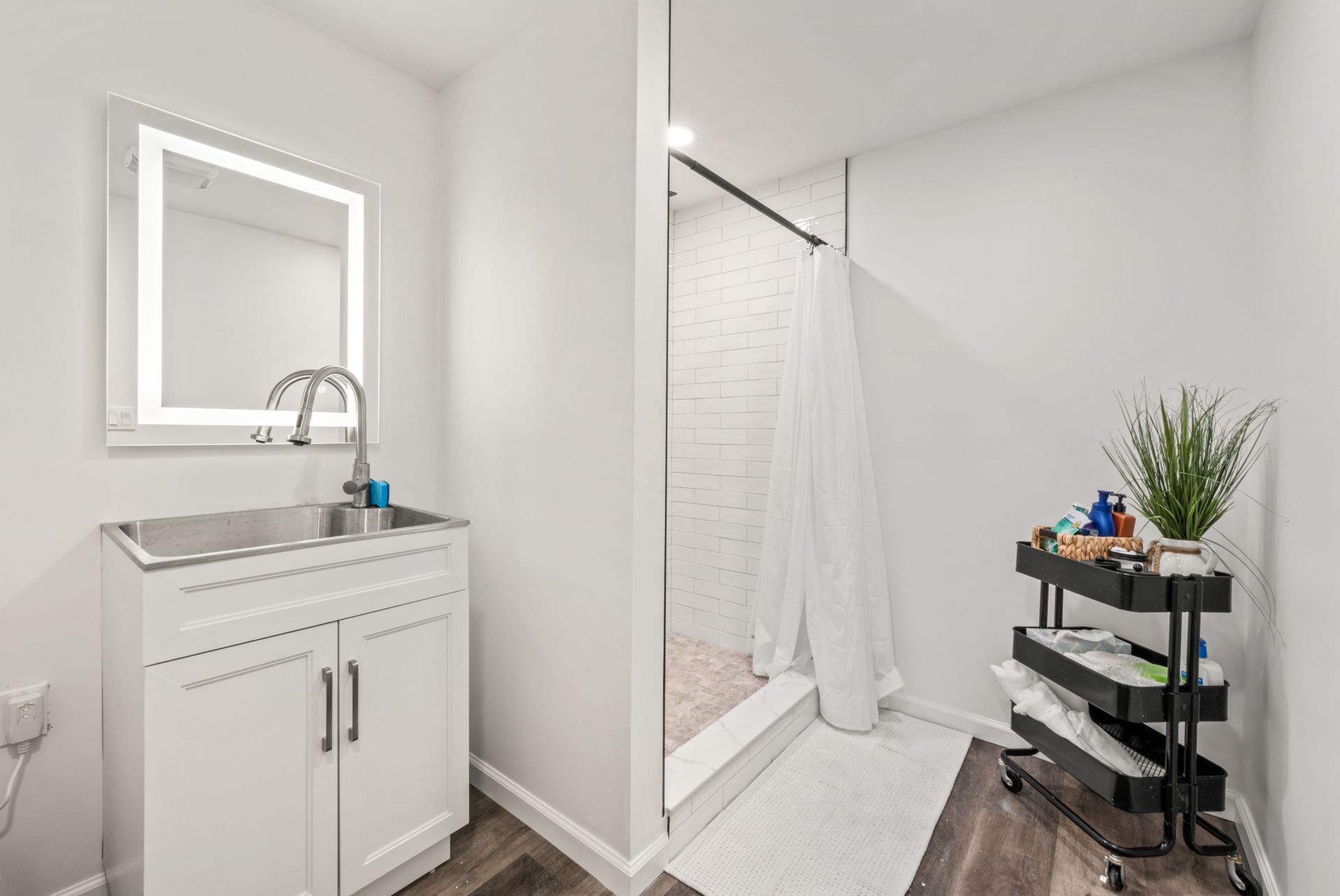 A modern, bright bathroom with a white vanity, a lit vanity mirror, a walk-in shower with white tile, and a black cart.