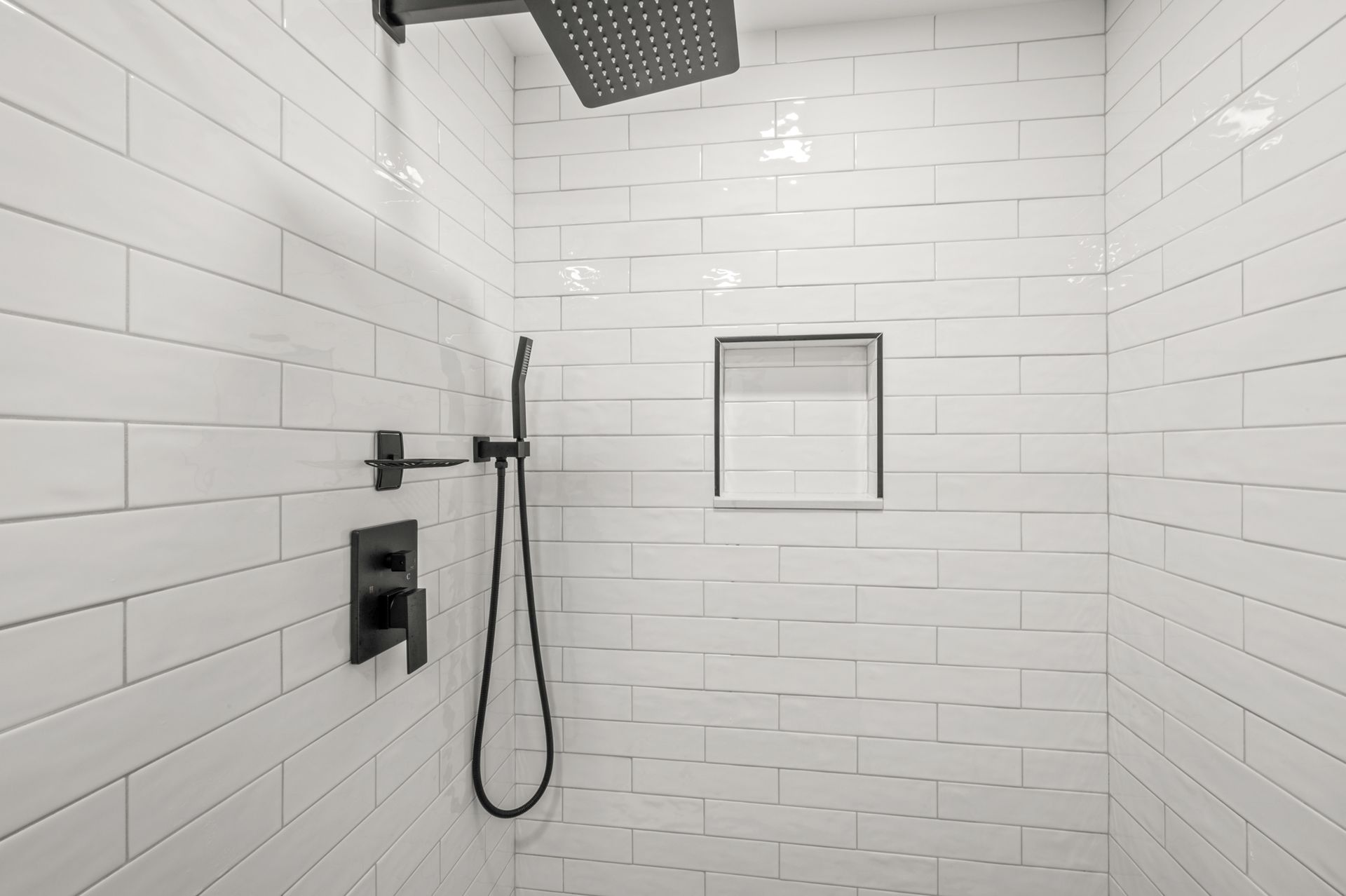 A walk-in shower with white subway tiles, matte black fixtures, a rainfall showerhead, a handheld sprayer, and a niche.