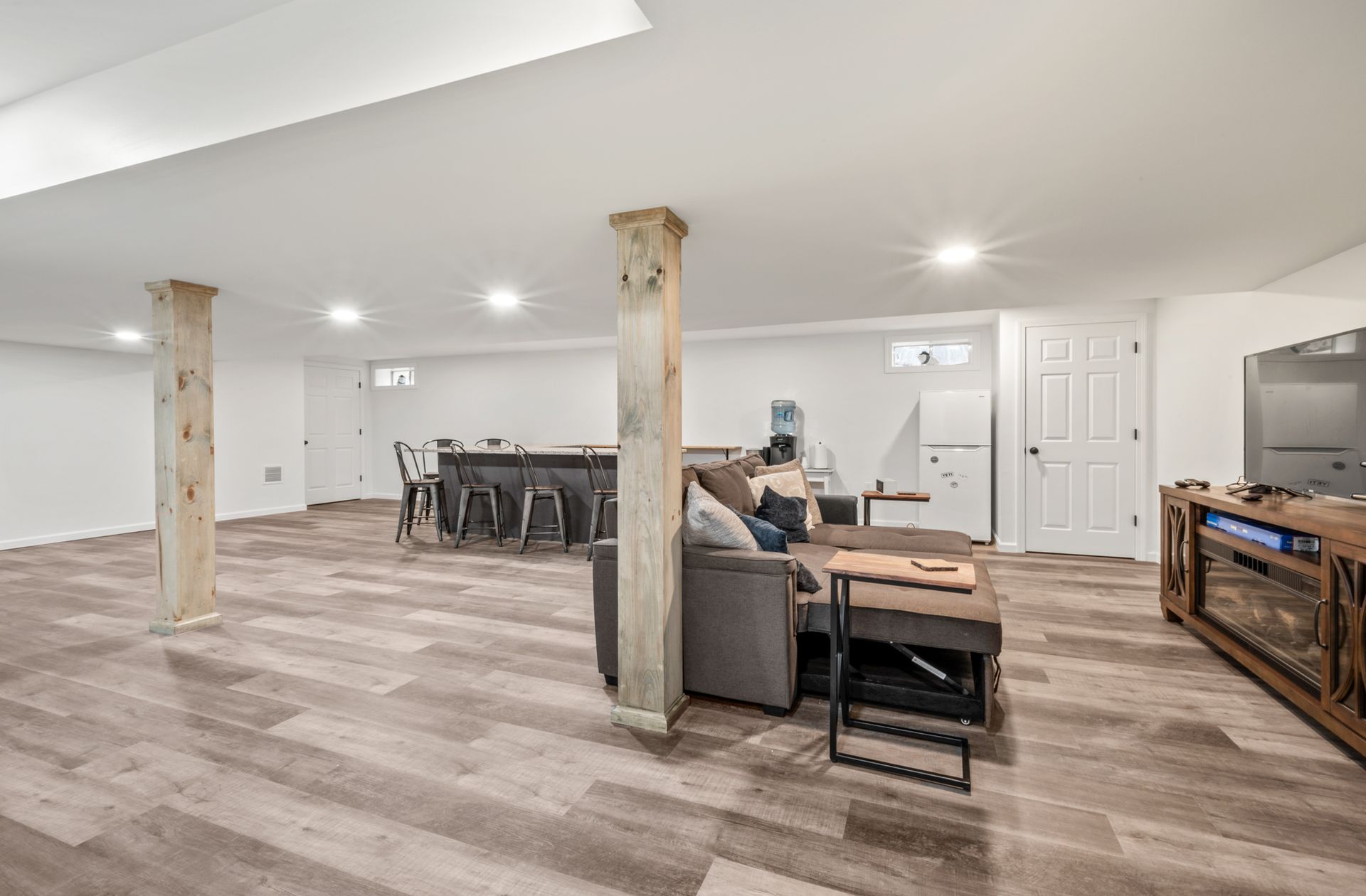 A modern basement featuring light wood floors, a sofa facing a TV console, and a bar area with stools behind wooden pillars.
