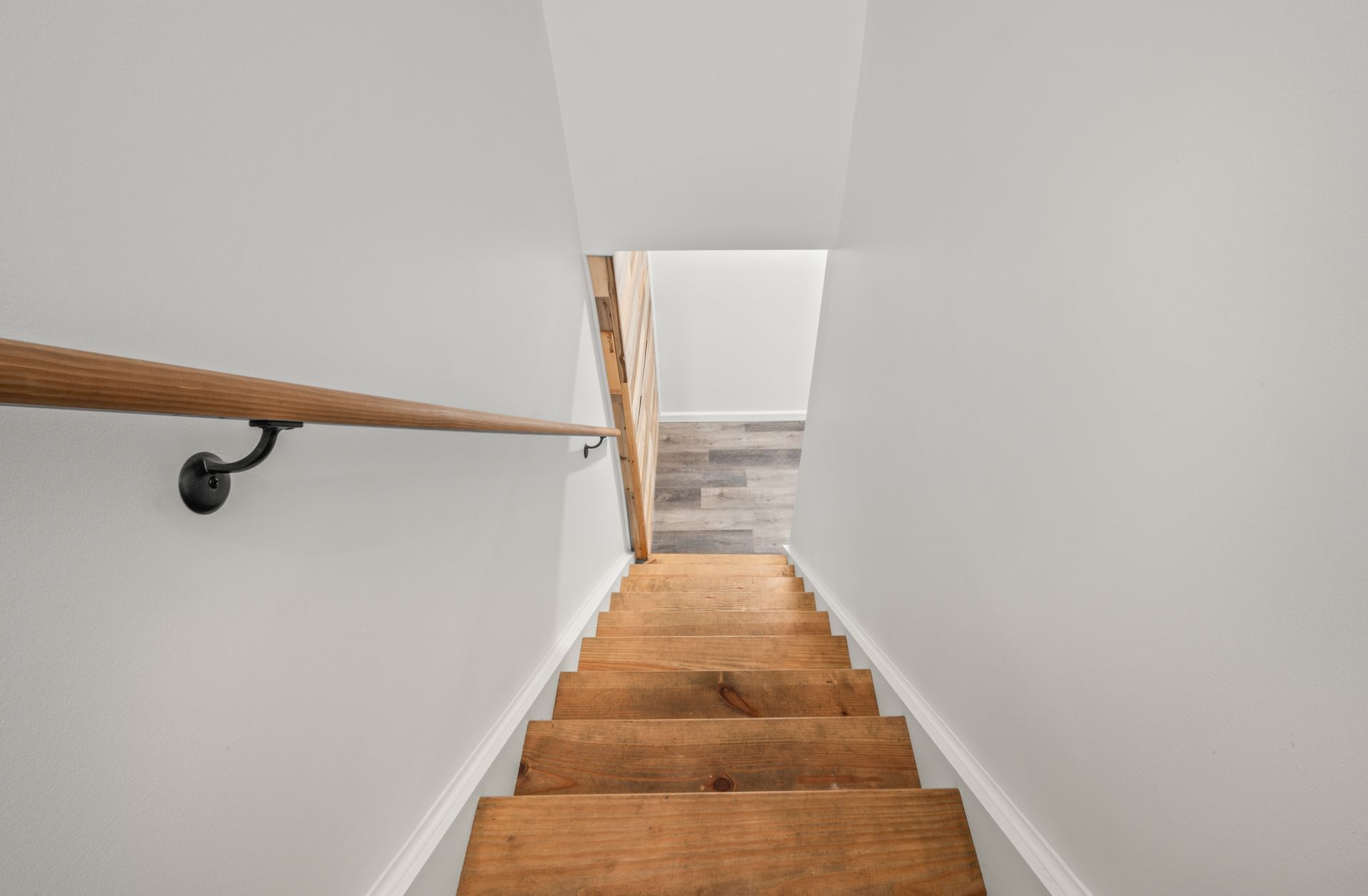A wooden staircase descends between plain white walls, featuring a simple wooden handrail mounted with black brackets.