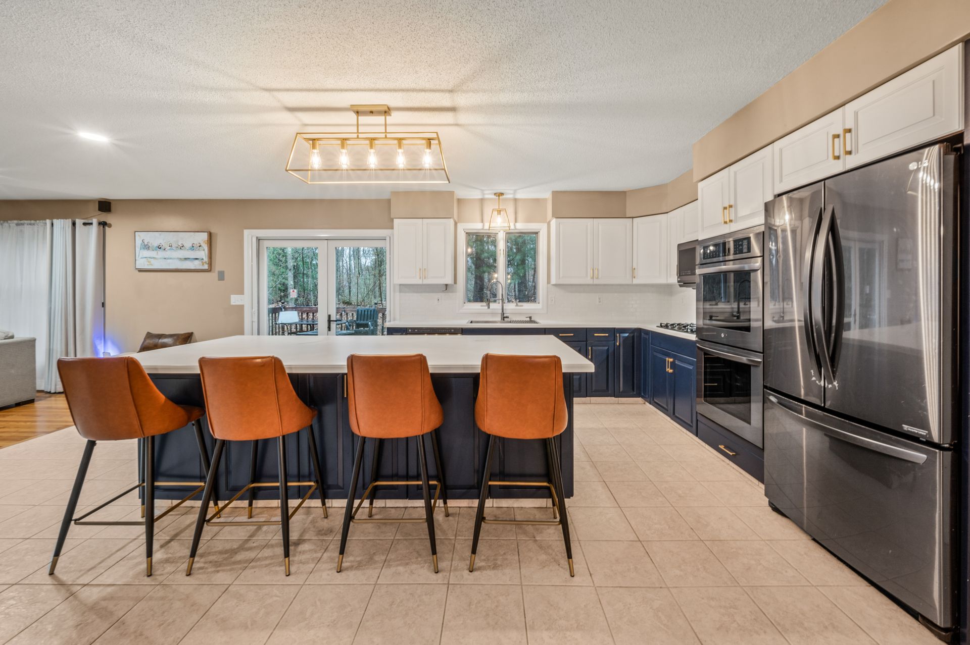 Modern kitchen with a large island, four tan bar stools, dark blue cabinets, white upper cabinets, and stainless fridge.