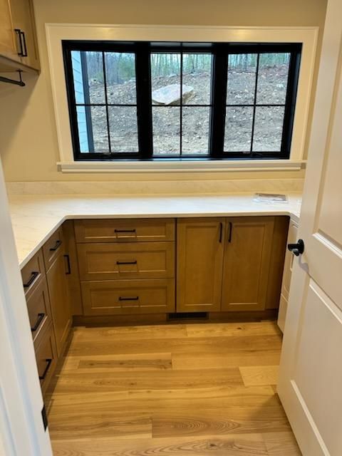 A laundry room with light wood cabinets, white countertops, dark window frames, and matching hardwood flooring.