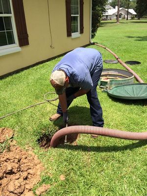 Person in blue shirt and jeans tending to a septic tank on a lawn, hose nearby.
