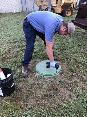 Person in blue shirt using a drill on a green lid in a grassy area; a bucket and construction vehicle are nearby.