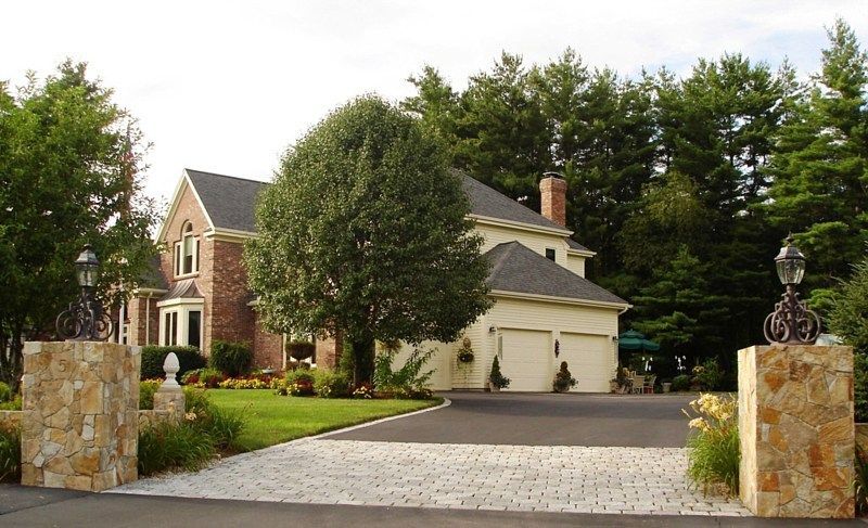 A two-story brick and siding house with a paved driveway between two stone pillars topped with lanterns.