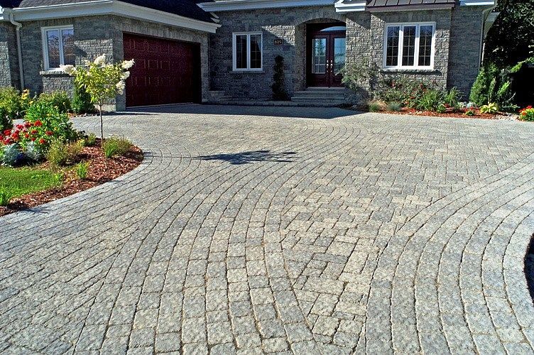 A stone paver driveway with a curved, patterned layout leading to the entrance of a grey stone residential house.
