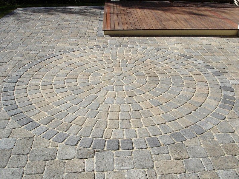 A circular pattern of gray and tan paving stones set within a larger patio next to a wooden deck.