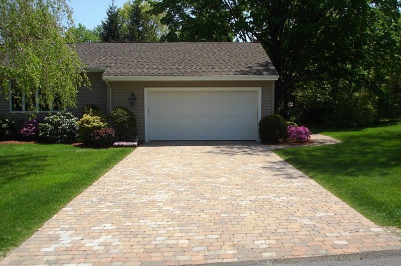A wide, paved driveway made of multi-colored stone pavers leading to a white, two-car garage at a suburban home.
