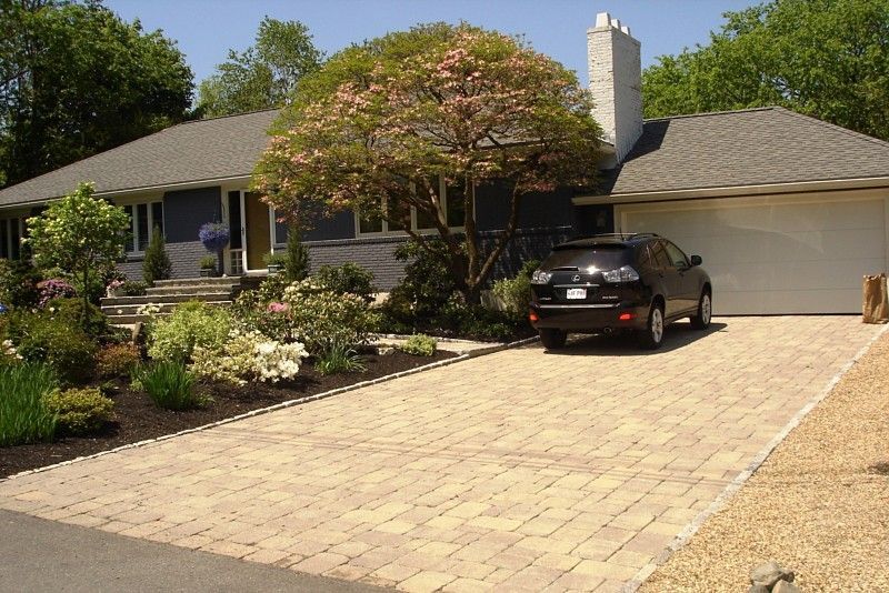 A one-story suburban home with a paved stone driveway, a dark car, and a flowering tree in the front yard.