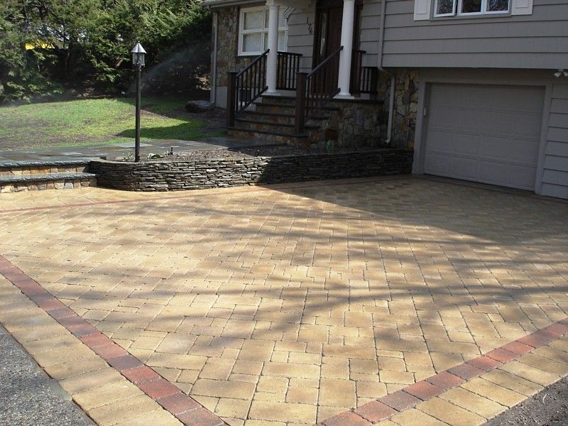 A tan paver driveway with a red border leads to the stone-walled entrance and garage of a gray-sided home.
