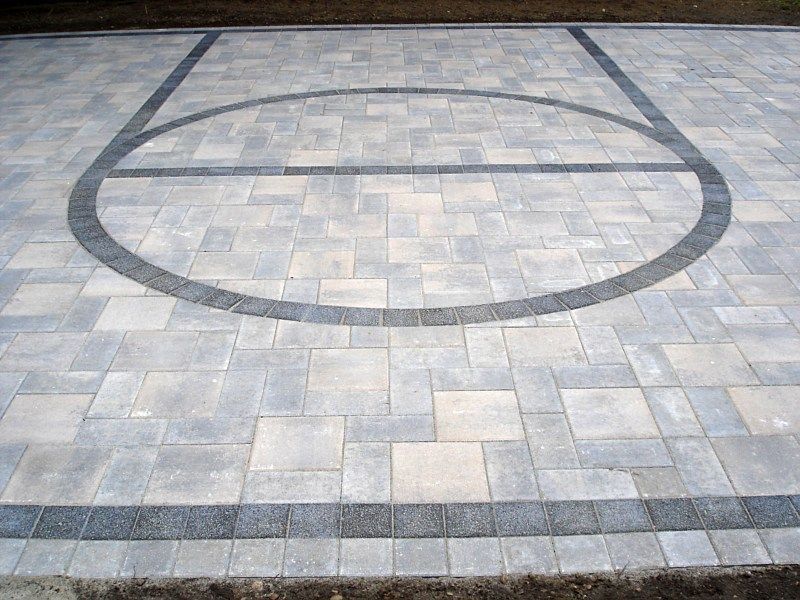 A basketball court surface made of light gray paving stones with a darker gray stone inlay forming the court markings.