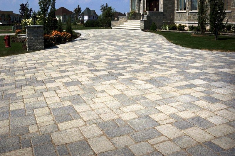 A paved driveway with multi-sized, interlocking gray and beige stone pavers leading to a suburban home entrance.