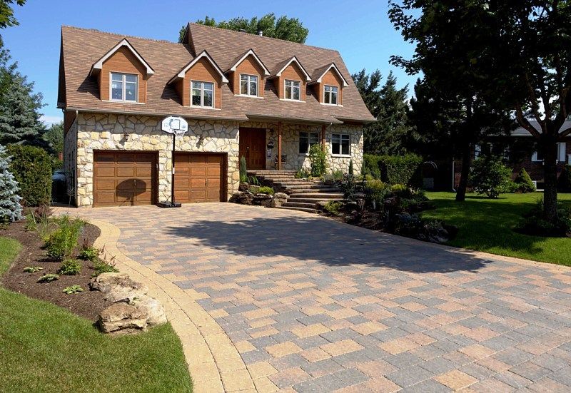 A two-story stone house with a brown roof and a large, multi-toned paved driveway in a suburban setting.