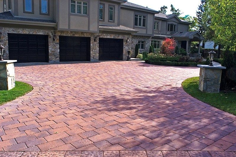A large house with a multi-car garage fronted by a wide, patterned red brick driveway and two stone entrance pillars.