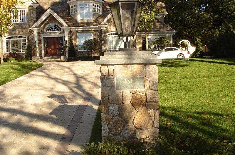 A stone pillar with a lantern stands in the foreground of a large suburban house with a paved driveway and green lawn.
