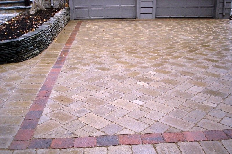 A paved driveway featuring tan-colored interlocking bricks bordered by a distinct row of dark red rectangular stones.