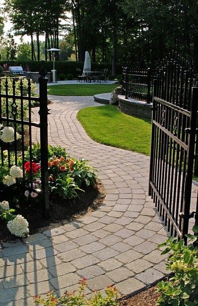 A curved stone paver walkway leads through an open black metal gate toward a landscaped backyard patio area.