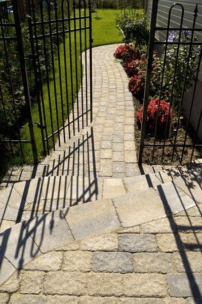 A stone walkway leads through an open metal gate towards a patch of green grass and flowering shrubs.