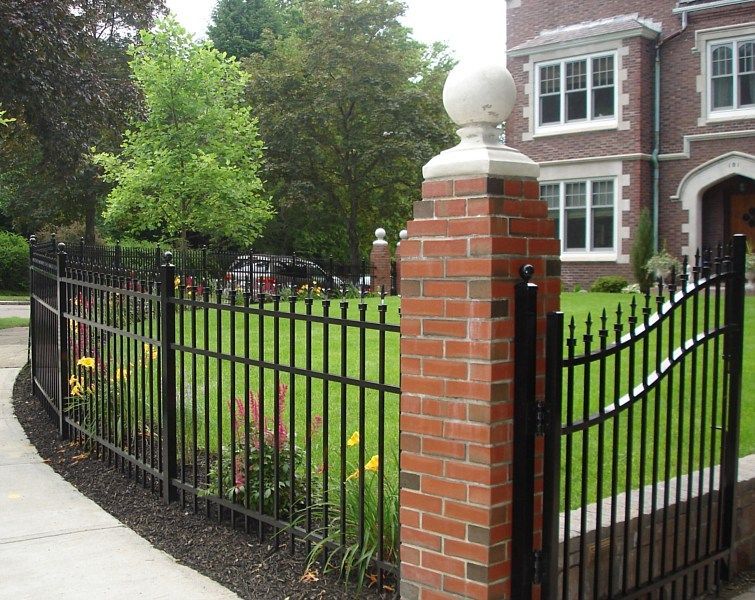 A red brick gate pillar topped with a white sphere, connecting black wrought iron fencing in front of a brick home.
