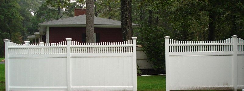 A white vinyl privacy fence with a decorative picket top stands in front of a house in a wooded area.