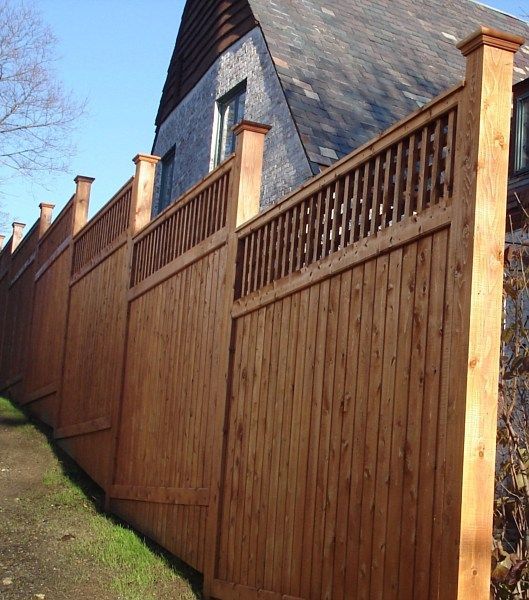 A tall, natural wood fence with a lattice top section runs along a grassy path in front of a house with a shingled roof.